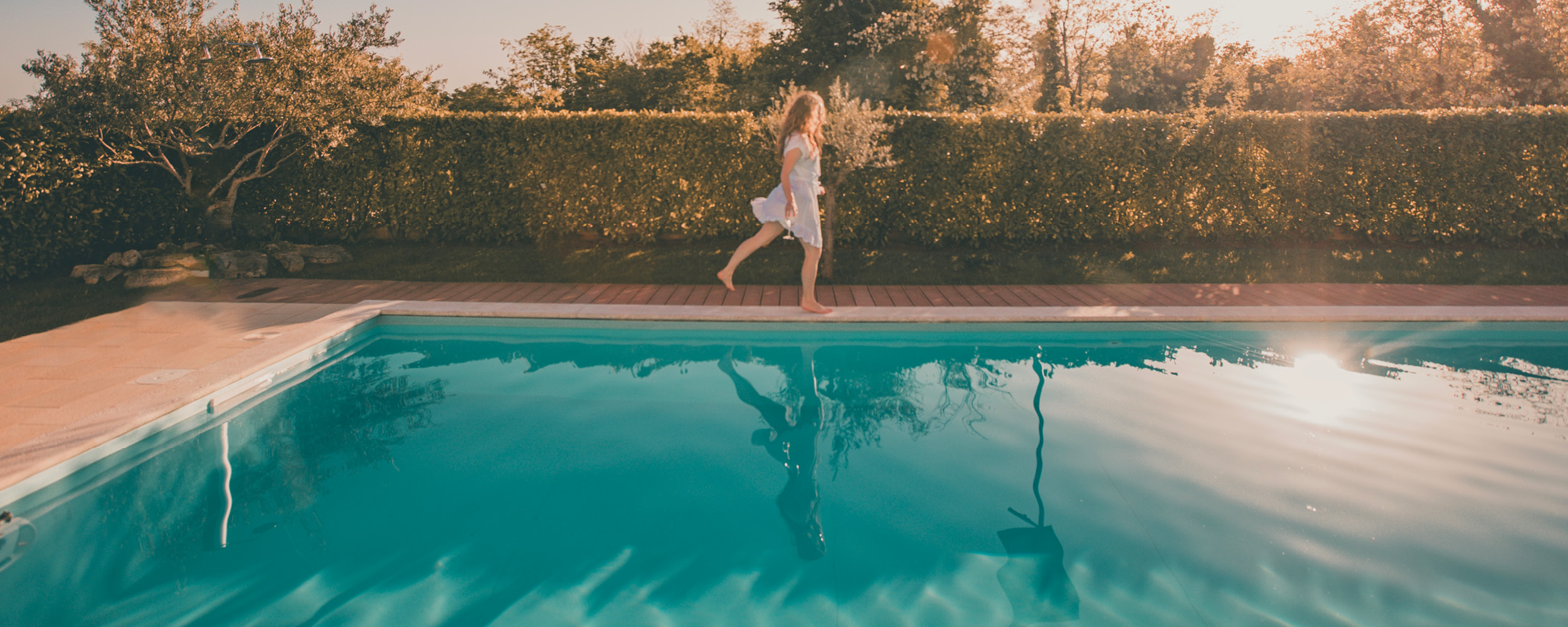 Scène estivale relaxante autour d'une piscine coque avec eau turquoise et bords en carrelage beige, entourée d'une haie verdoyante sous une lumière dorée, illustrant confort et qualité de vie.