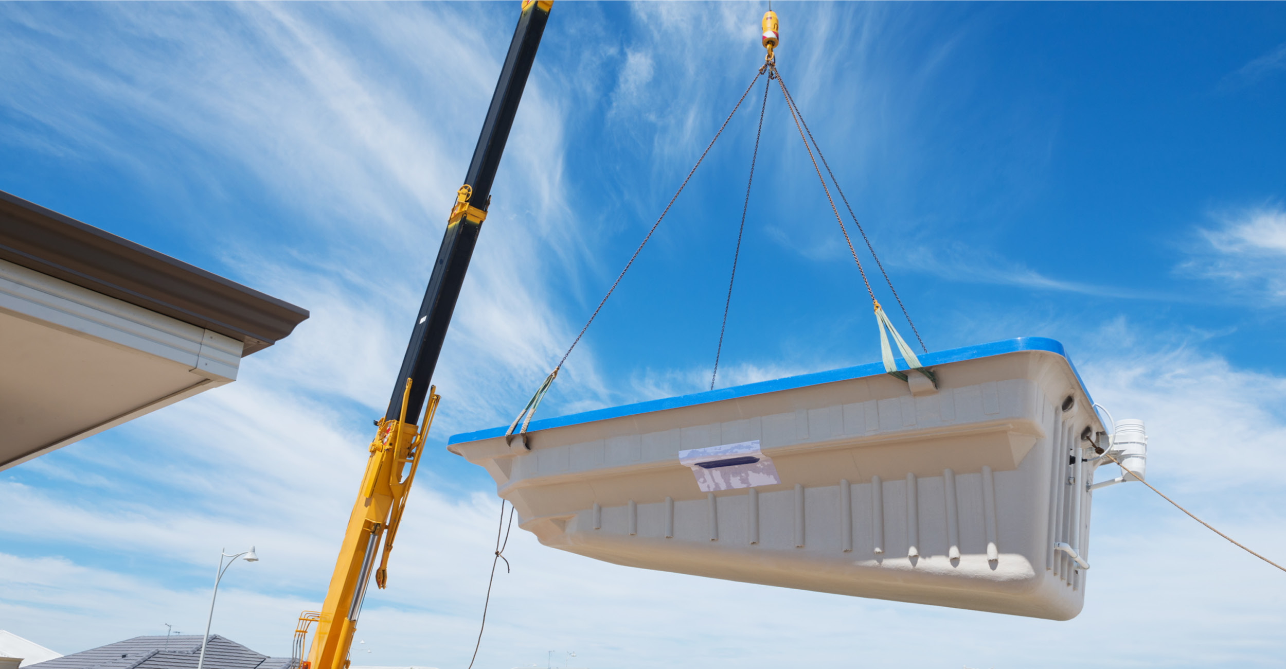 Installation professionnelle de piscine coque en polyester suspendue par une grue, journée ensoleillée en zone résidentielle.