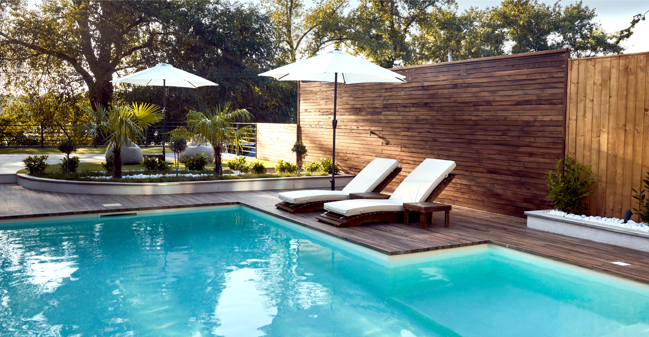 Espace extérieur élégant autour d'une piscine coque turquoise, avec chaises longues blanches, parasols, terrasses en bois, végétation tropicale, et éléments décoratifs, créant une ambiance de luxe et de détente.