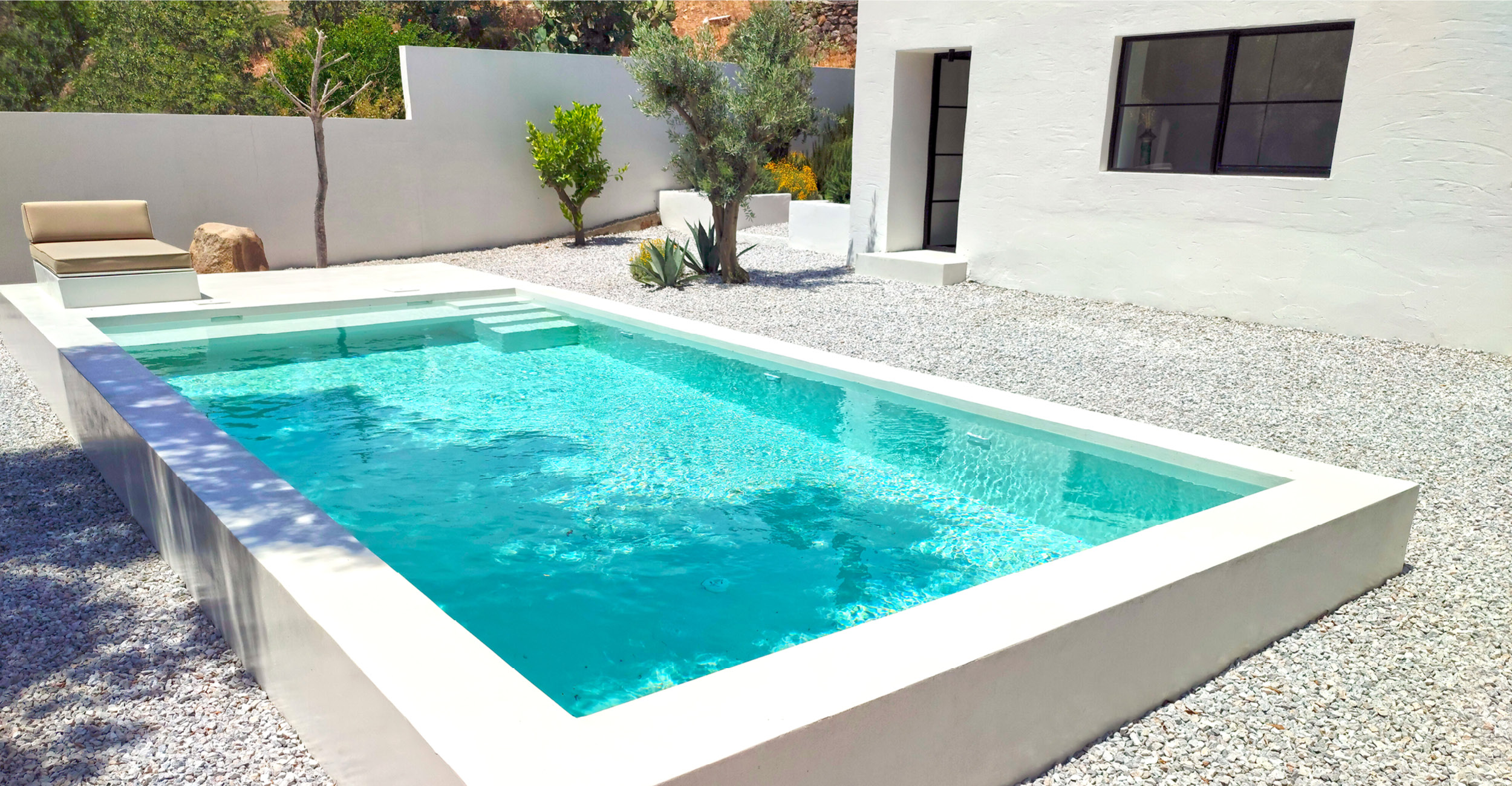 Piscine coque moderne rectangulaire avec eau turquoise entourée de gravier clair et végétation succulente, accompagnée d'un bain de soleil beige, devant une maison épurée aux murs blancs et grandes fenêtres, créant un espace extérieur contemporain et relaxant