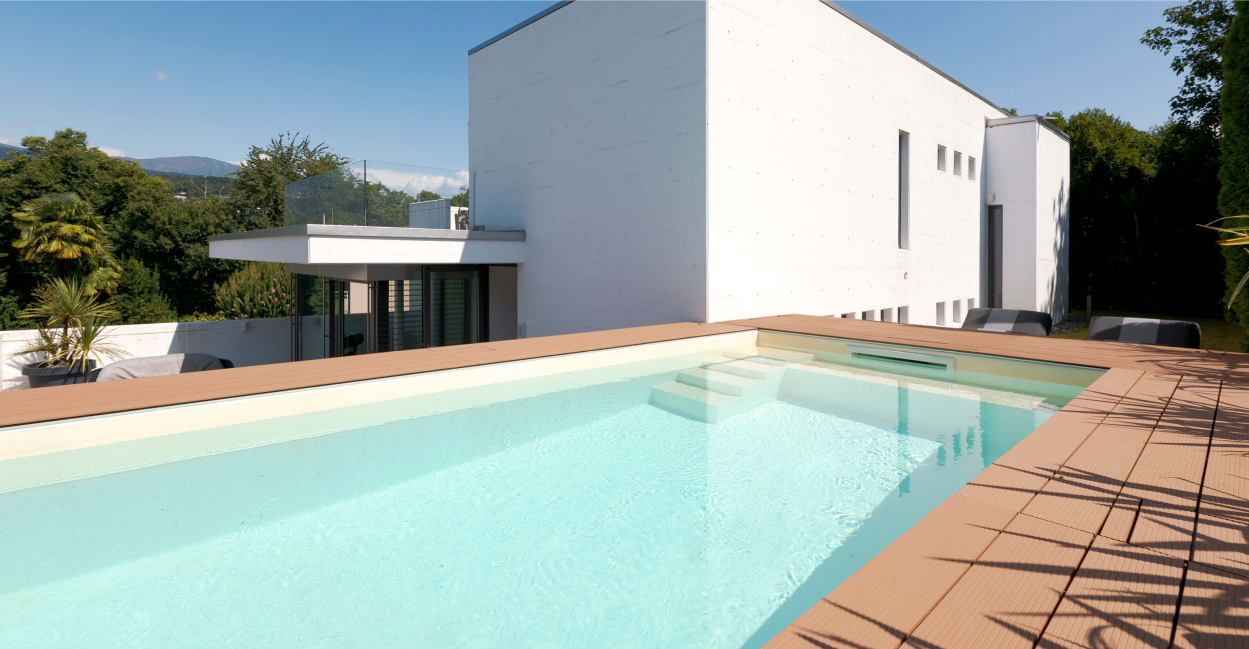 Piscine coque élégante avec eau bleu clair, entourée de lames de bois, intégrée dans un environnement moderne avec maison contemporaine et palmiers, créant une ambiance relaxante et esthétique.