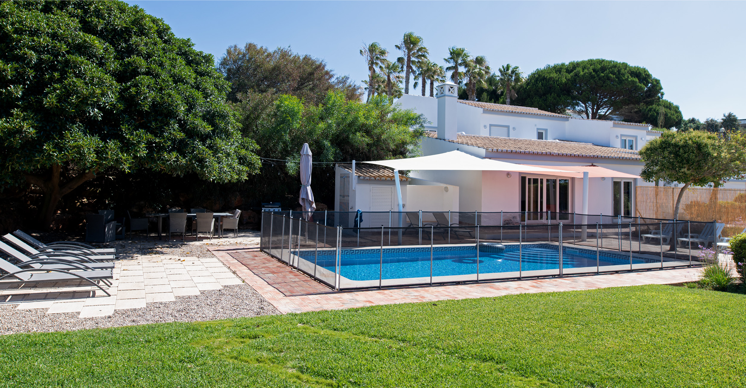 Jardin avec piscine coque entourée de sécurité en verre, pelouse verte, mobilier d'extérieur avec chaises longues et table, maisons modernes avec grandes fenêtres, ambiance détente et loisirs.