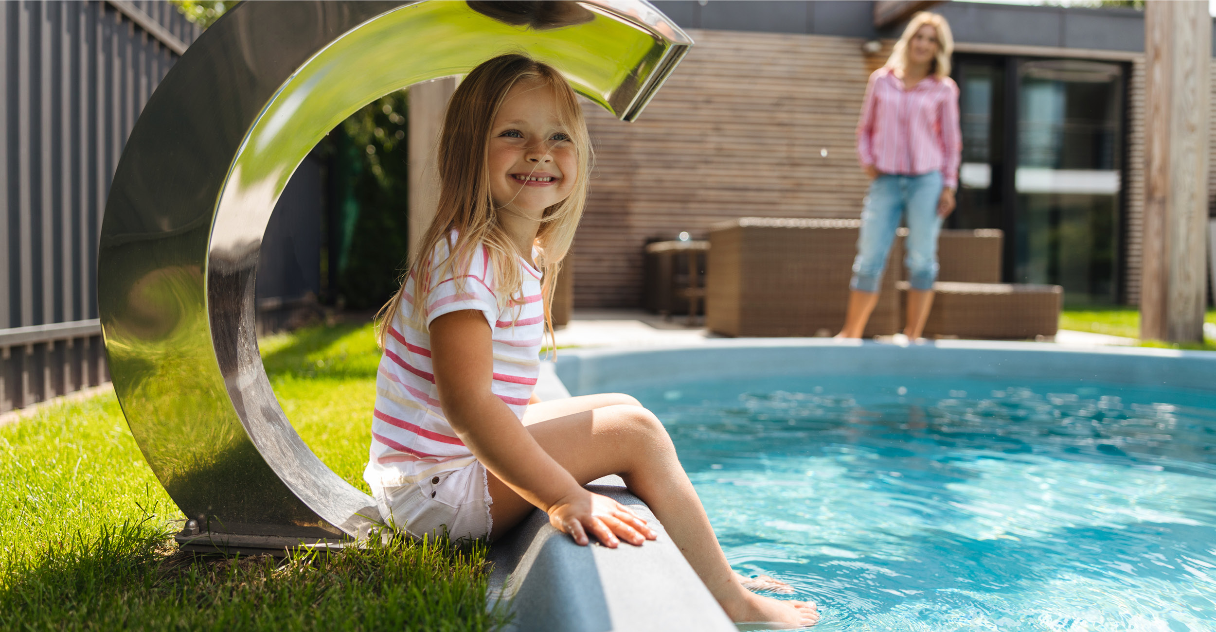 Piscine coque ovale avec jet d'eau et coin salon familial, entourée de gazon vert, scène avec enfant souriant pour une ambiance conviviale et moderne.