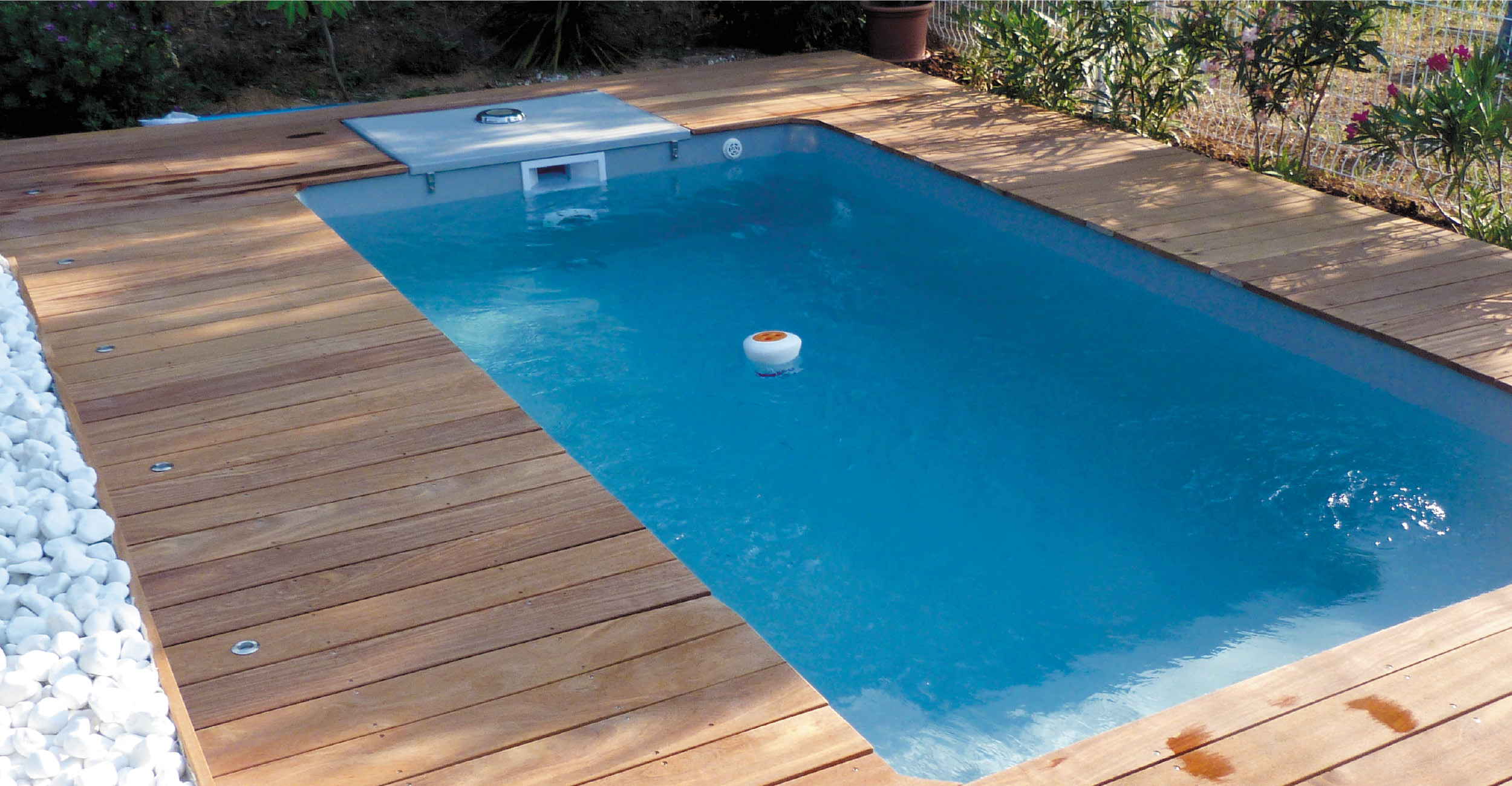 Piscine coque moderne rectangulaire avec bords en bois, galets blancs et végétation luxuriante, équipée de skimmer et flotteur pour entretien optimal