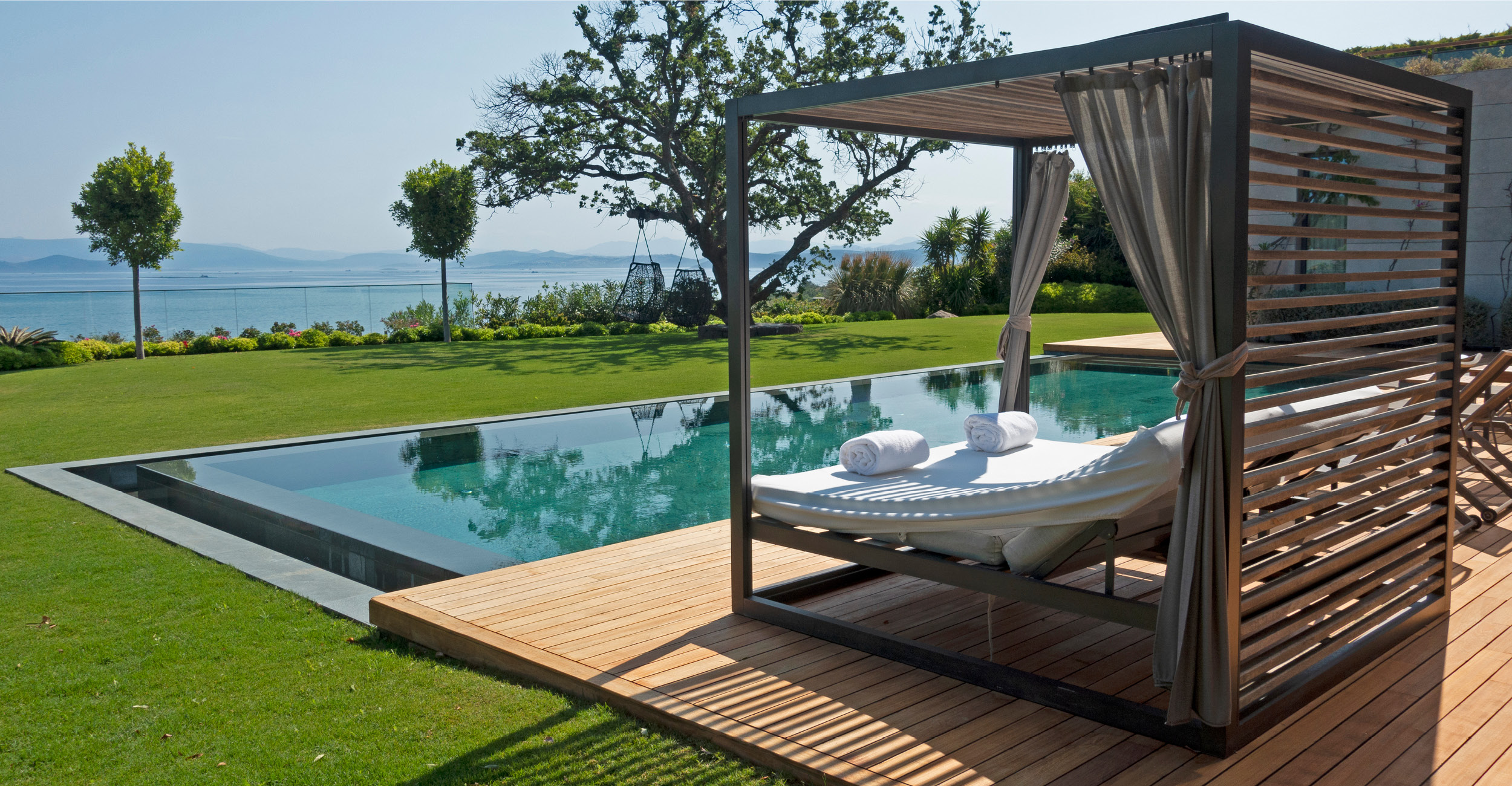 Piscine coque à débordement moderne avec espace détente en bois, pelouse verdoyante et plantes ornementales, reflétant luxe et confort pour un extérieur chic.