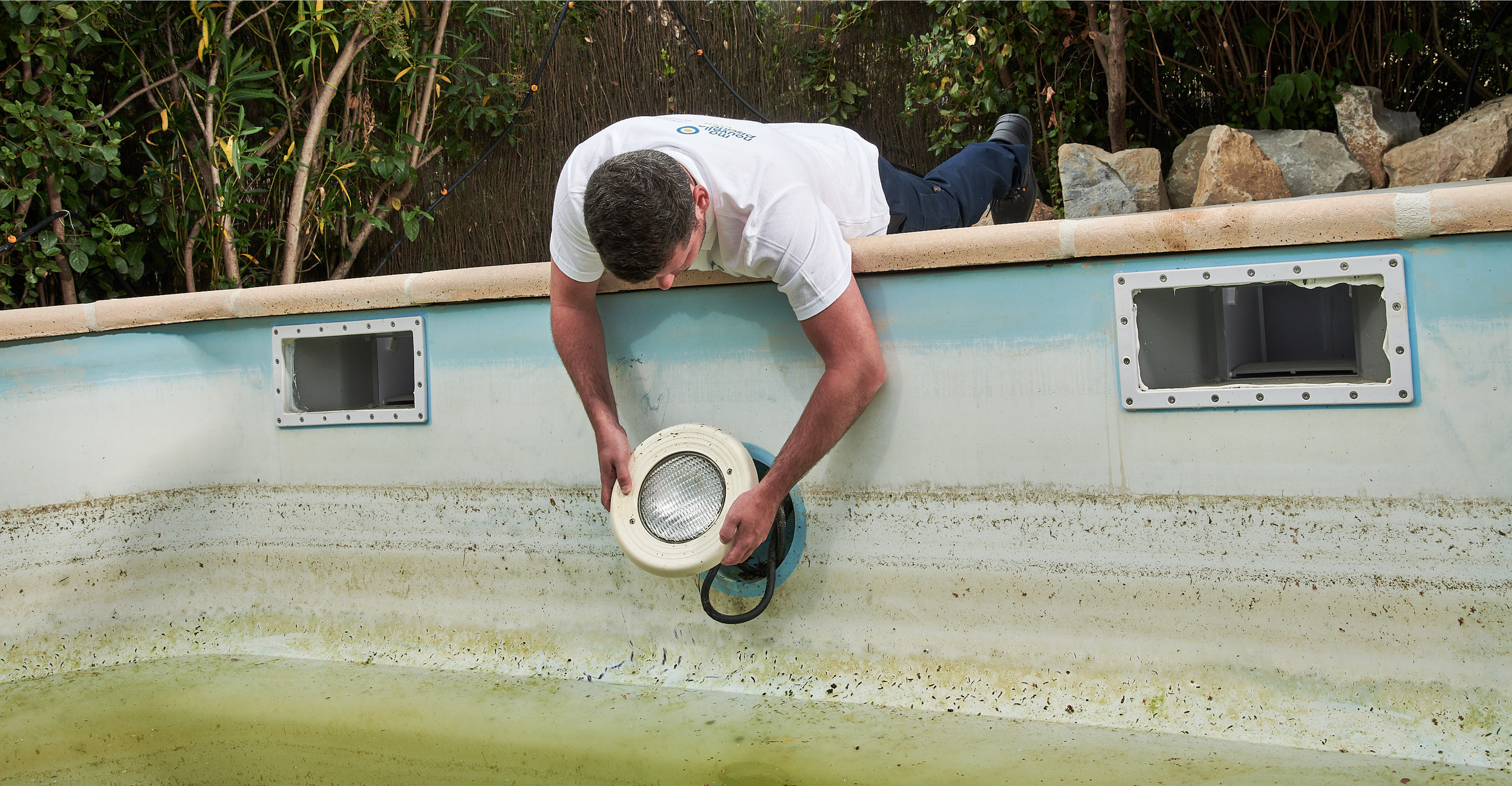 Technicien vérifiant une lumière de piscine coque pour maintenance dans un environnement naturel avec signes d'usure et végétation environnante