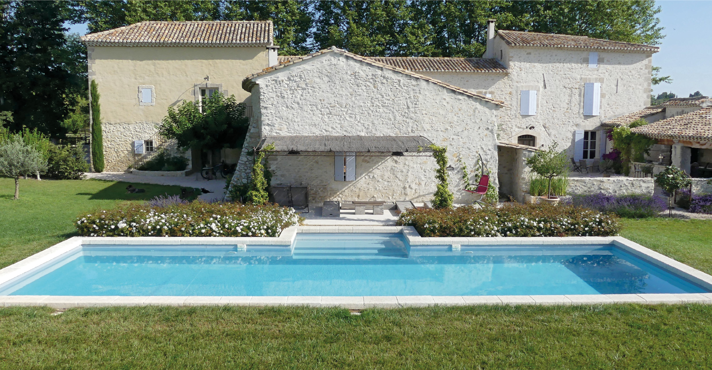 Piscine coque rectangulaire aux eaux claires entourée de margelles en pierre, nichée dans un jardin verdoyant avec des fleurs colorées et des oliviers, près de maisons en pierre avec toit en tuiles, créant une ambiance paisible et naturelle.