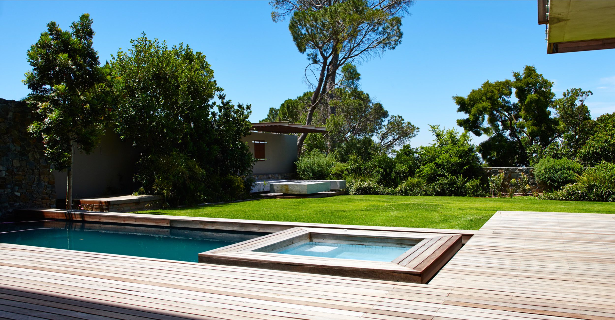 Espace extérieur aménagé avec piscine coque rectangulaire moderne et spa, entouré de terrasse en bois et jardin verdoyant avec cabanon en arrière-plan, sous un ciel dégagé pour un cadre luxueux et serein.