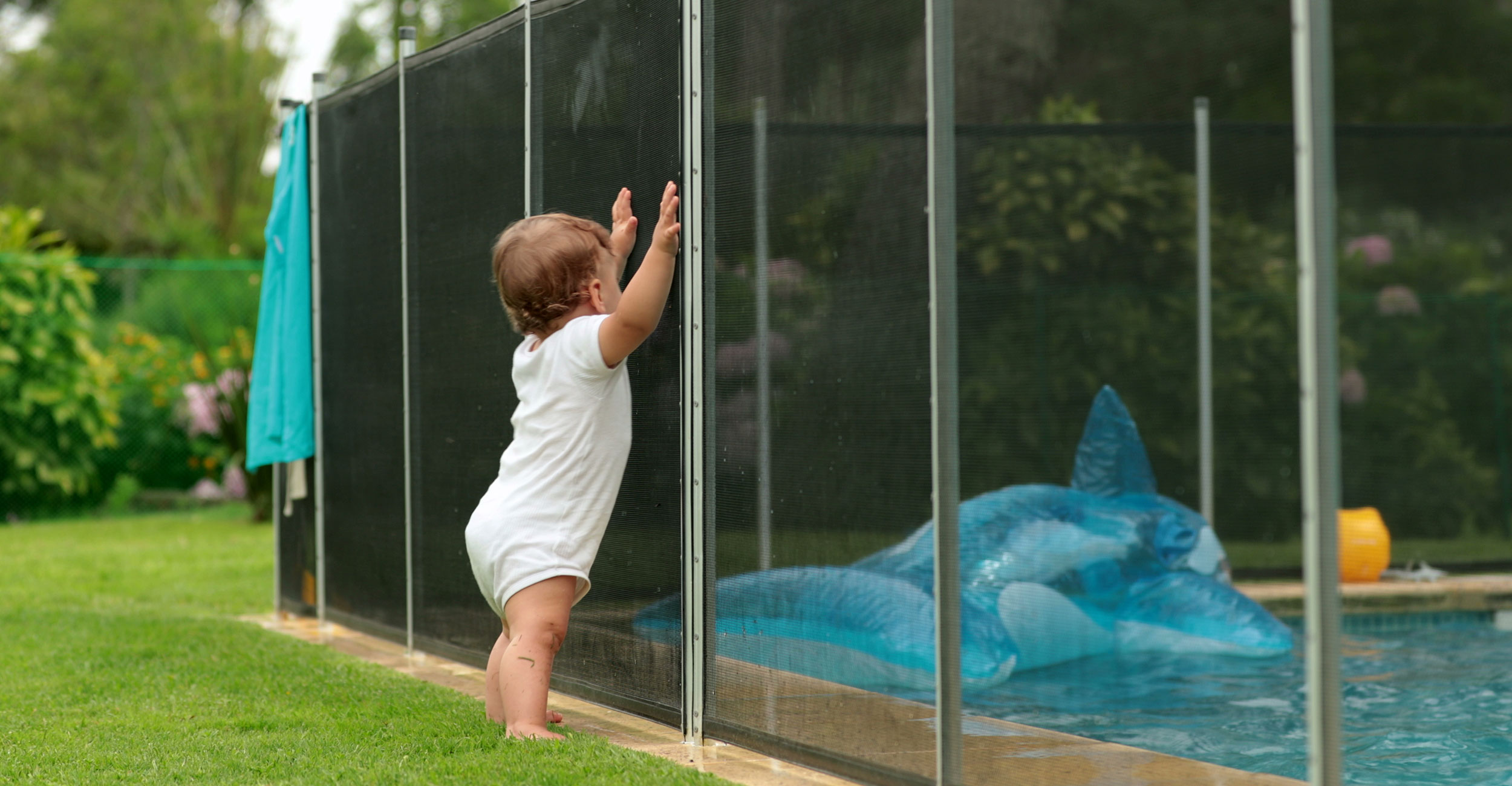 Scène familiale apaisante autour d'une piscine coque sécurisée avec clôture, bébé en tenue blanche à proximité, grand jouet gonflable requin, environnement naturel verdoyant, soulignant sécurité et plaisir estival pour enfants et familles.