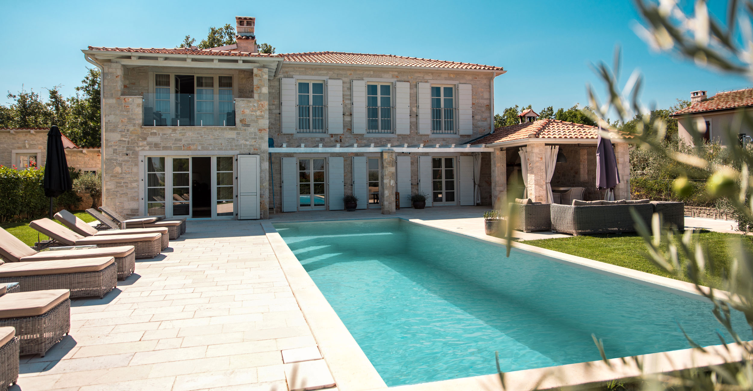 Piscine coque rectangulaire dans un cadre résidentiel méditerranéen avec maison en pierre et volets bleus, entourée de chaises longues et verdure, reflétant luxe et détente