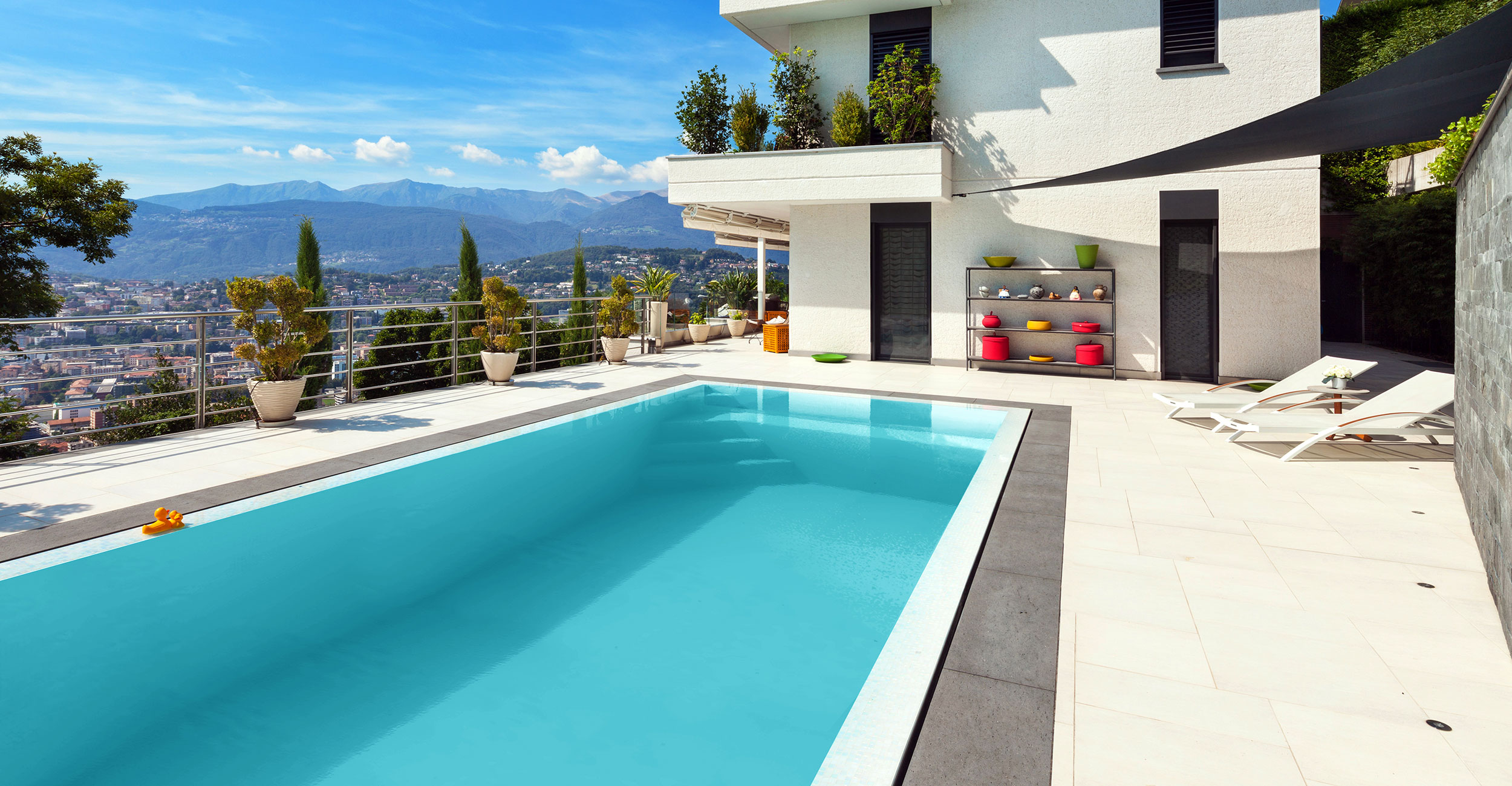 Piscine coque moderne intégrée dans un cadre urbain avec vue sur les montagnes, eau bleue claire, canard en plastique flottant, entourée de chaises longues et plantes sur terrasse en pierre, maison contemporaine en arrière-plan