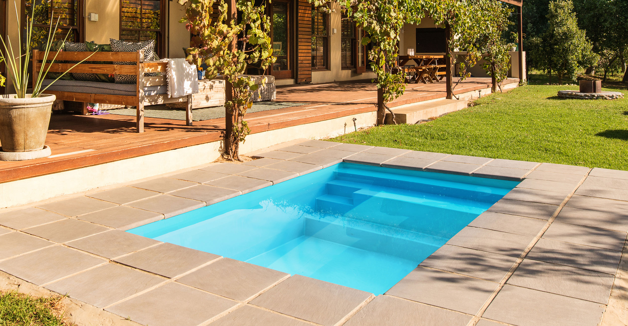 Piscine coque moderne avec eau bleue éclatante et contour en pierre grise, entourée de mobilier extérieur confortable et végétation luxuriante, idéale pour un espace détente et convivial.