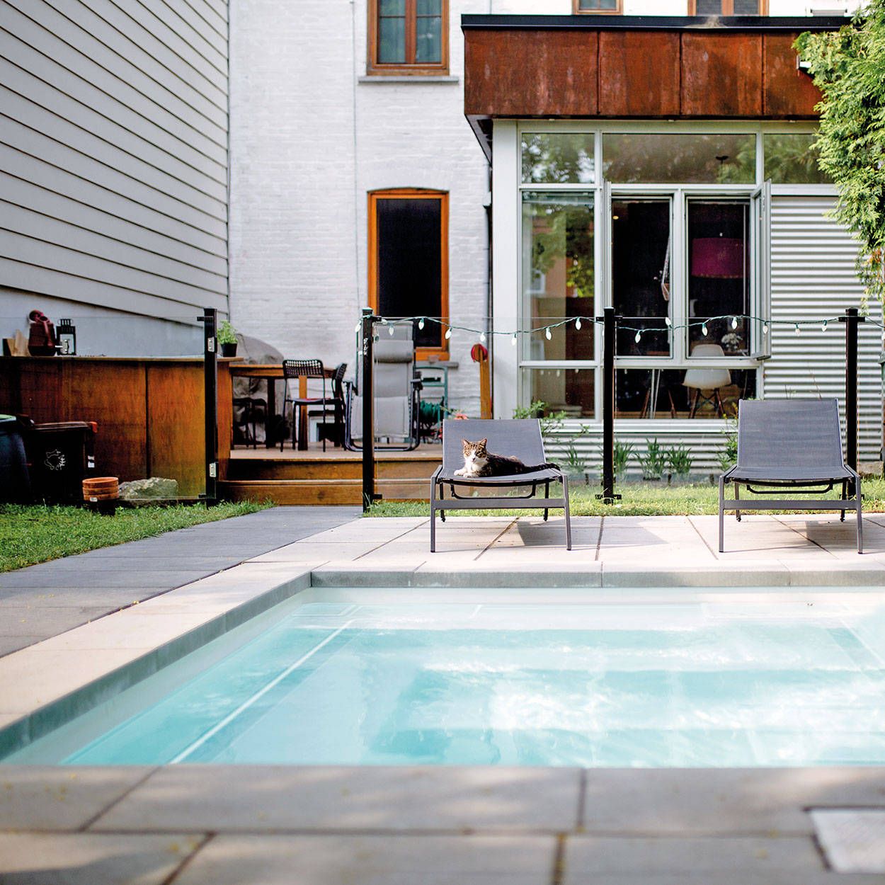 Espace extérieur moderne avec piscine coque rectangulaire, entourée de chaises longues noires, terrasse aménagée et jardin verdoyant, éclairé naturellement, création de détente pour maisons contemporaines.