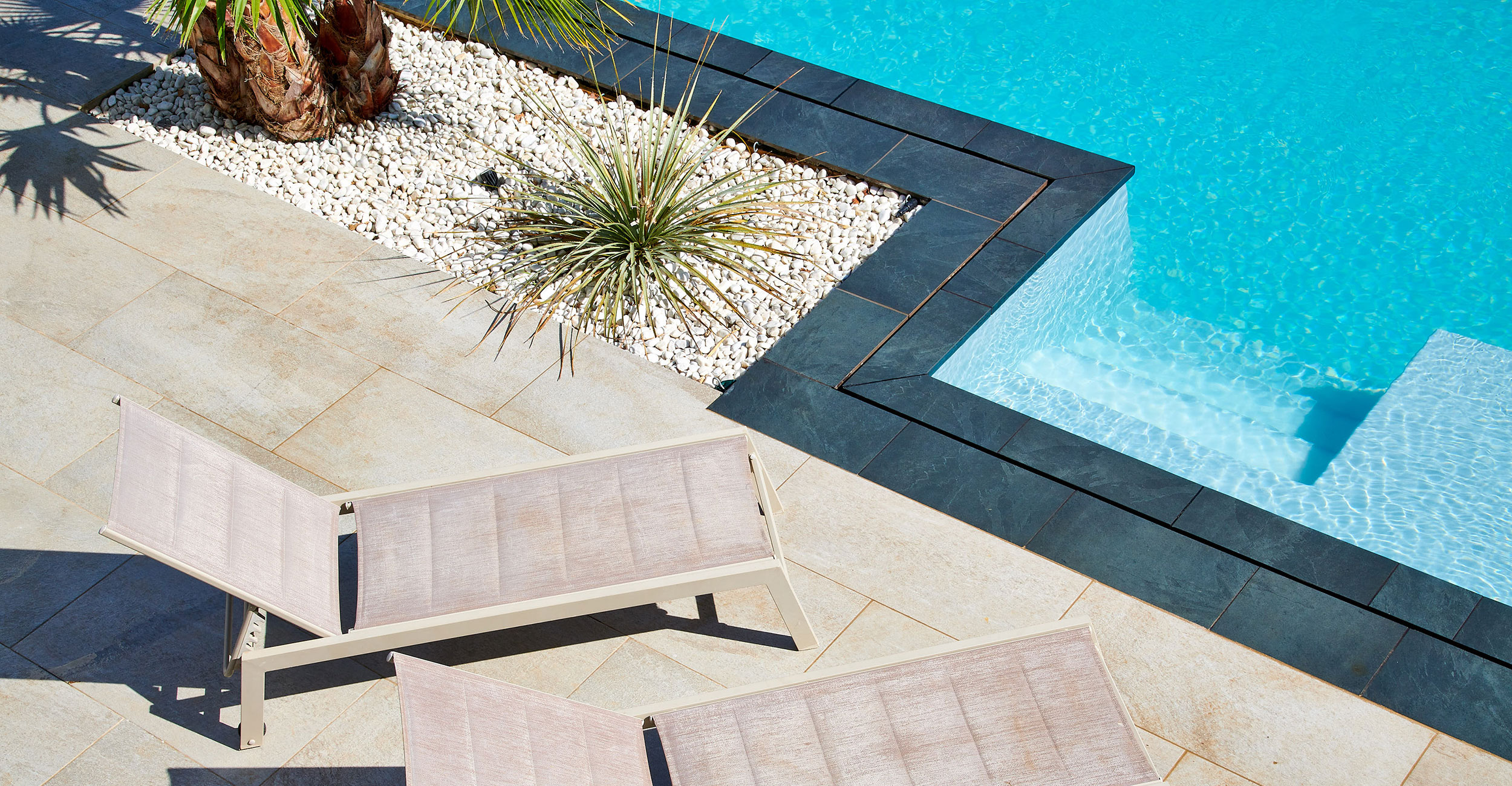 Espace extérieur moderne avec piscine coque bleue, terrasse en dalles neutres, chaises longues design, pierre blanche, et aménagement paysager tropical avec palmiers et succulentes.