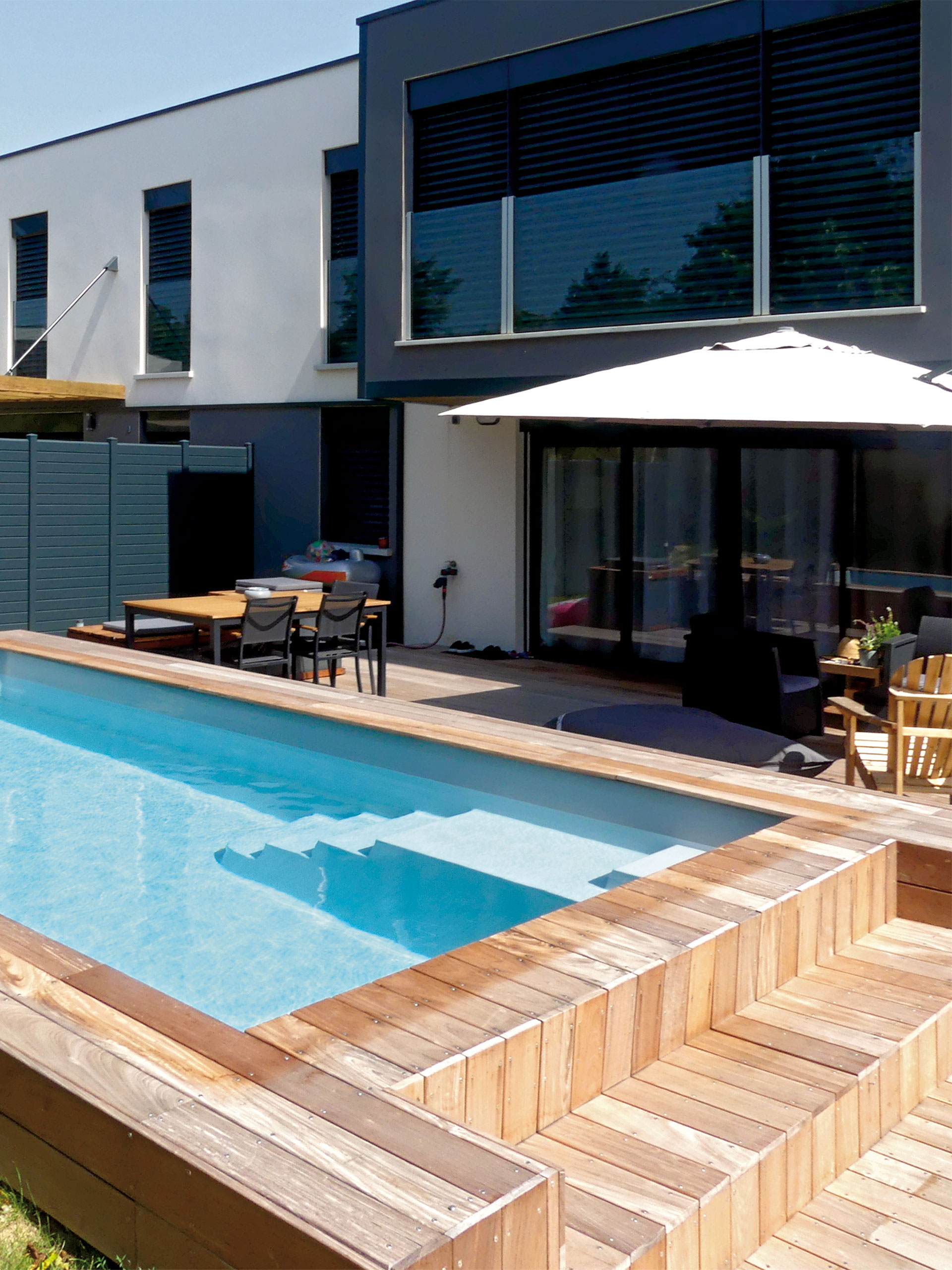 Piscine coque moderne avec terrasse en bois et mobilier de jardin, entourée de verdure, intégrée à une architecture contemporaine